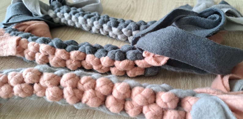 Three colorful braided dog tug toys in shades of gray and peach laid on a light wooden surface.